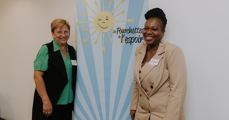 Deux femmes qui sourient devant l'affiche de l'organisme Les Fourchettes de l'Espoir